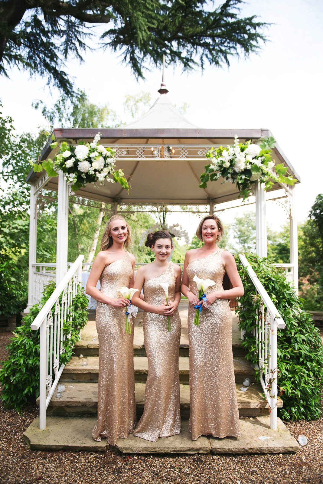 Bridesmaids wearing full length gold Champagne coloured sequin one shoulder bridesmaids dresses and carrying a single white lily.